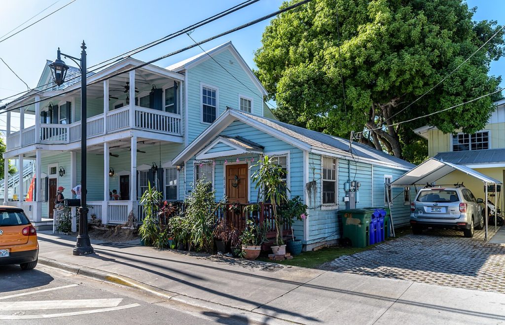 A light blue two-story house with porches and a small blue cottage next door on a sunny street.